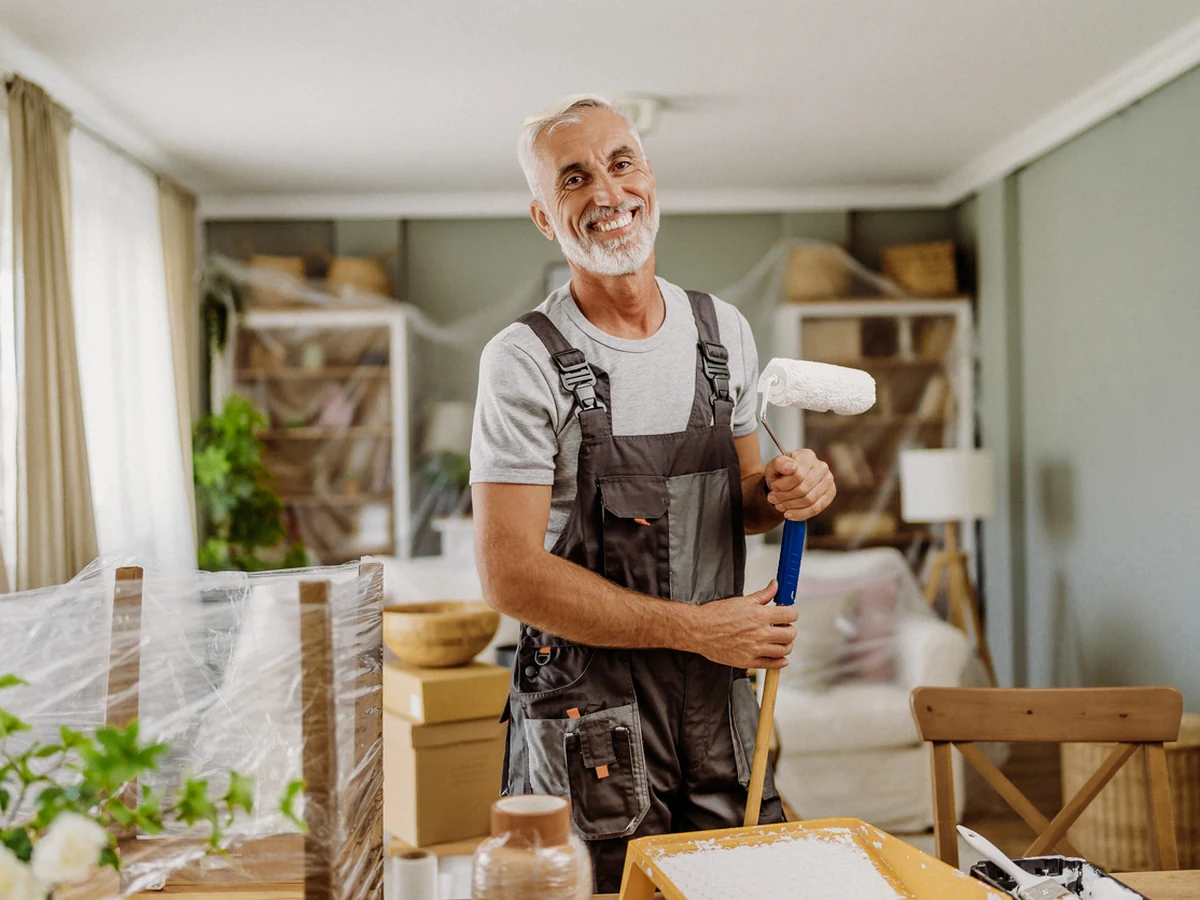 painting-decorating-older-man-holding-exeter-decorators Decorator holding a paint roller in a living room with the furniture covered in plastic sheeting